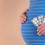 A close-up view of the belly of a pregnant woman in a blue T-shirt, which holds pills in her hands. The concept of medicine during pregnancy