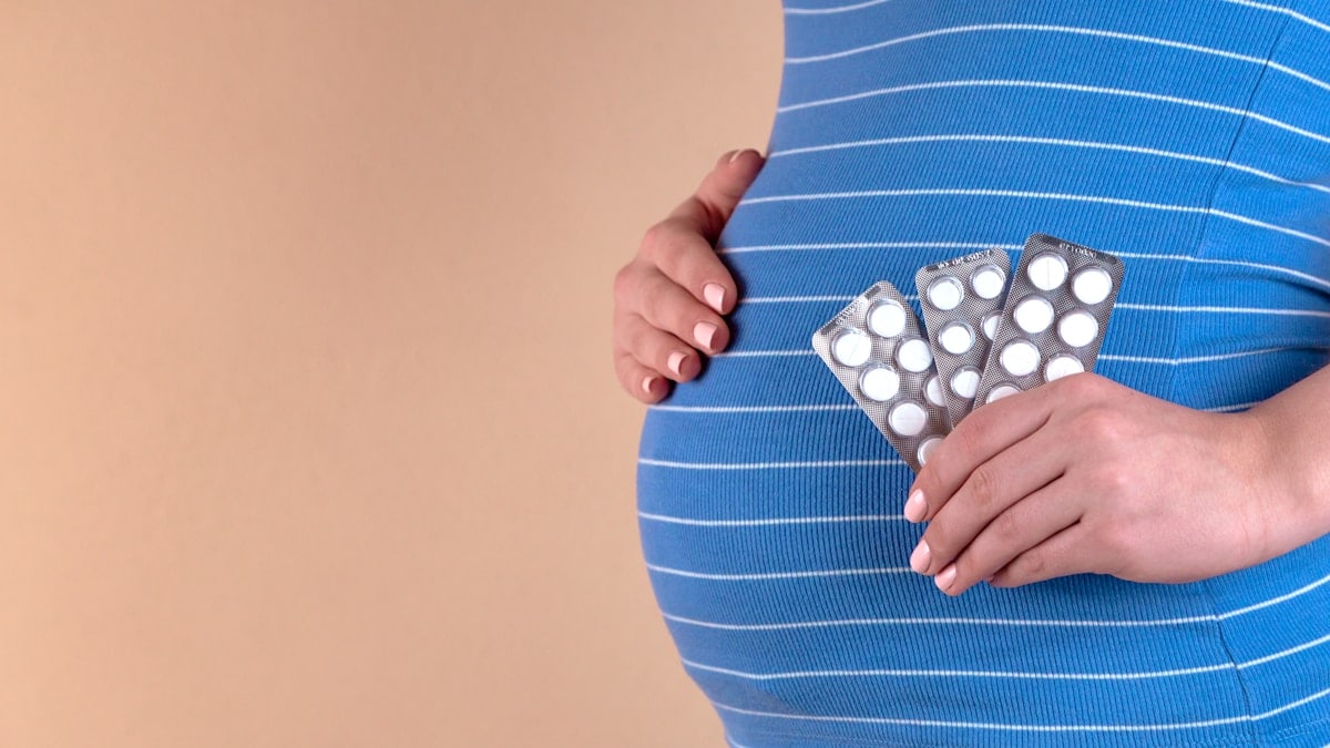 A close-up view of the belly of a pregnant woman in a blue T-shirt, which holds pills in her hands. The concept of medicine during pregnancy